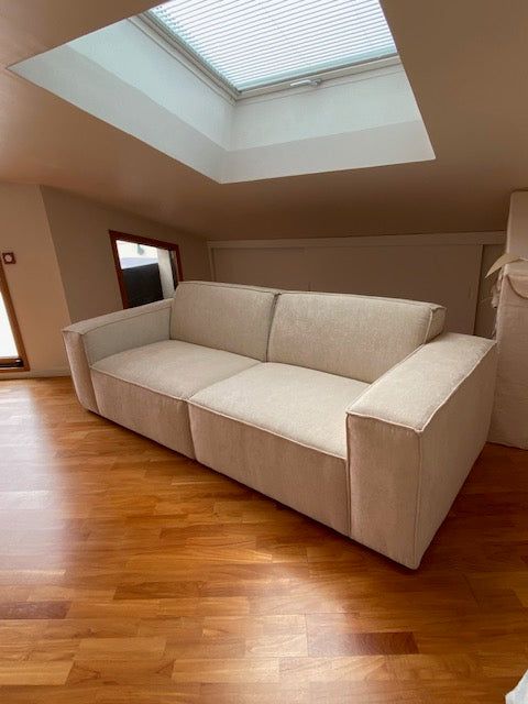 Retour client du canapé droit 4 places Gabrielle en tissu chenille beige apaisé de chez Paname Avenue en vue 3/4 dans un salon mansardé lumineux et minimaliste avec un parquet massif. 