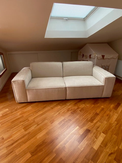 Retour client du canapé droit 4 places Gabrielle en tissu chenille beige apaisé de chez Paname Avenue en vue de face dans un salon lumineux et minimaliste avec un parquet massif. 