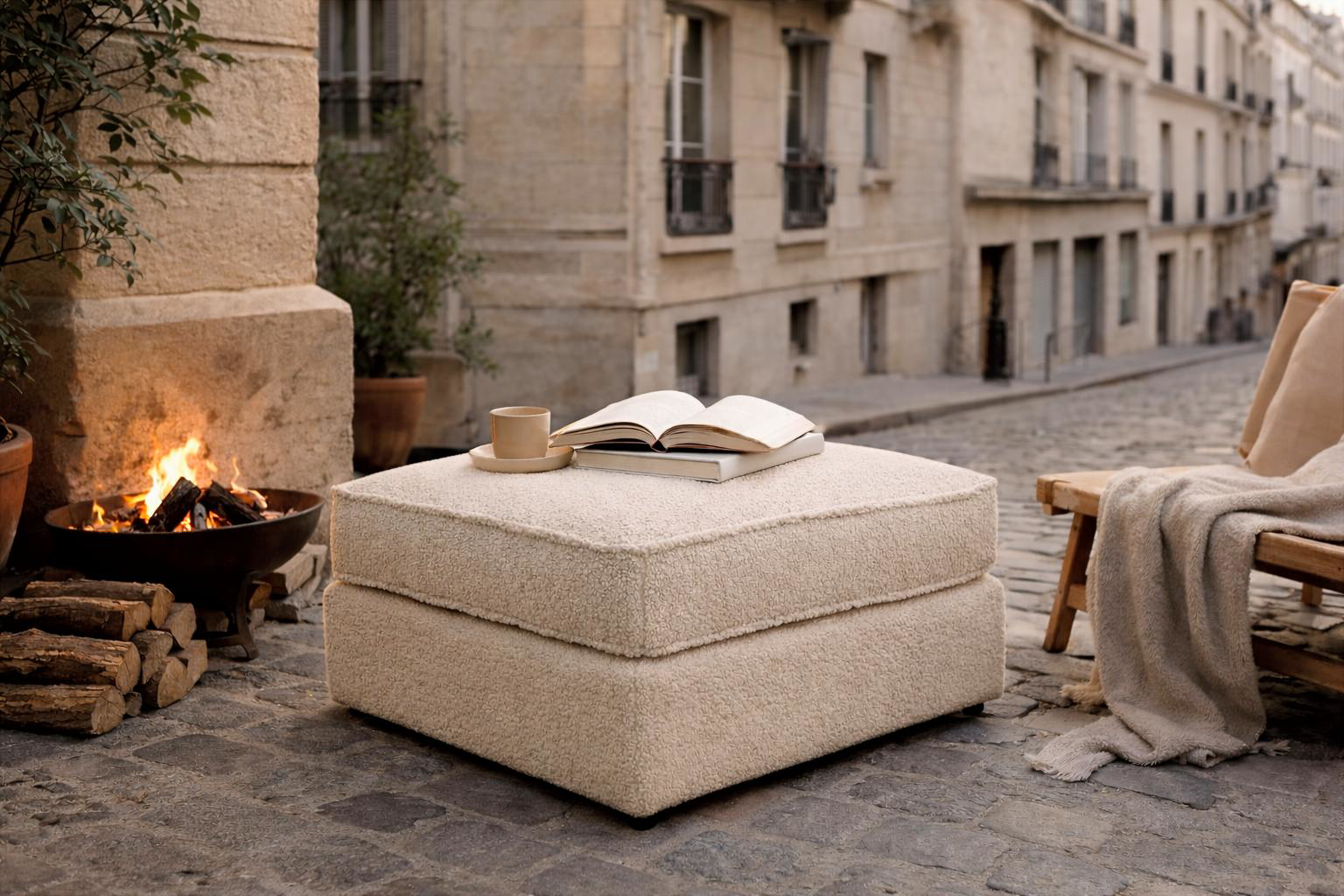 Pouf Durantin Paname Avenue en tissu bouclette beige utilisé en extérieur dans une rue pavée de Montmartre à Paris avec feu de bois, livre et ambiance chaleureuse