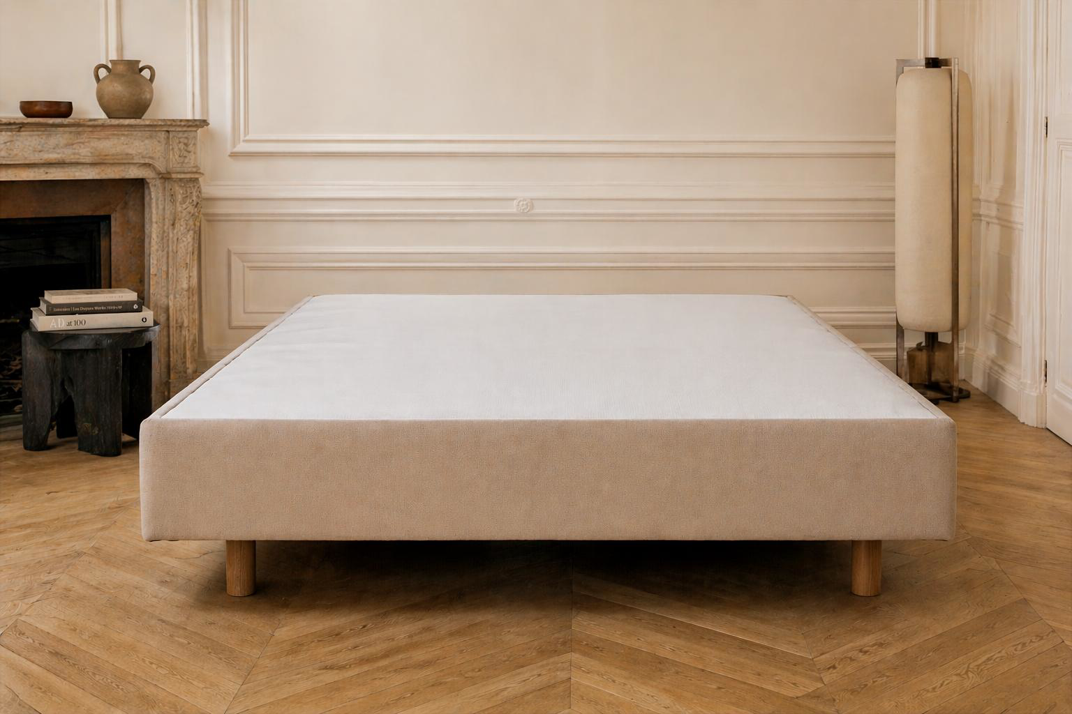 Sommier tapissier Richard Lenoir 140x200cm pour deux personnes, tissu beige et pieds bois, dans une chambre avec cheminée ancienne, moulures et luminaire colonne.