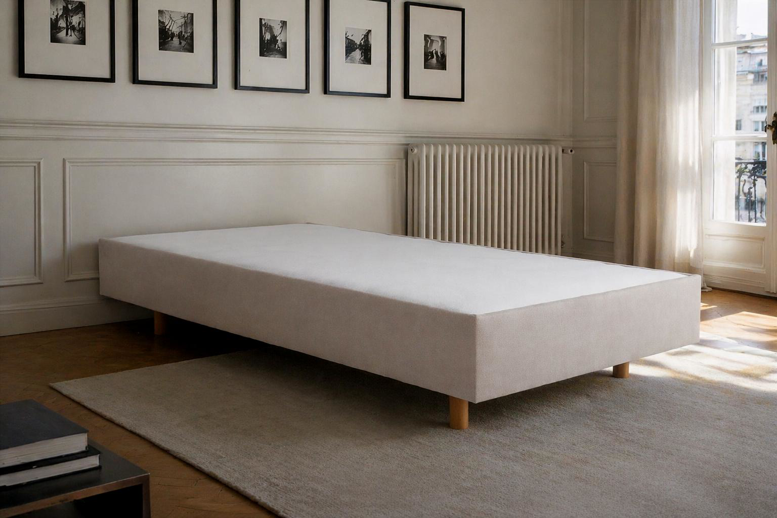 Sommier tapissier Richard Lenoir 80x200cm en tissu beige, vue de profil dans une chambre élégante avec parquet en chevrons, cadres muraux noirs et radiateur en fonte.